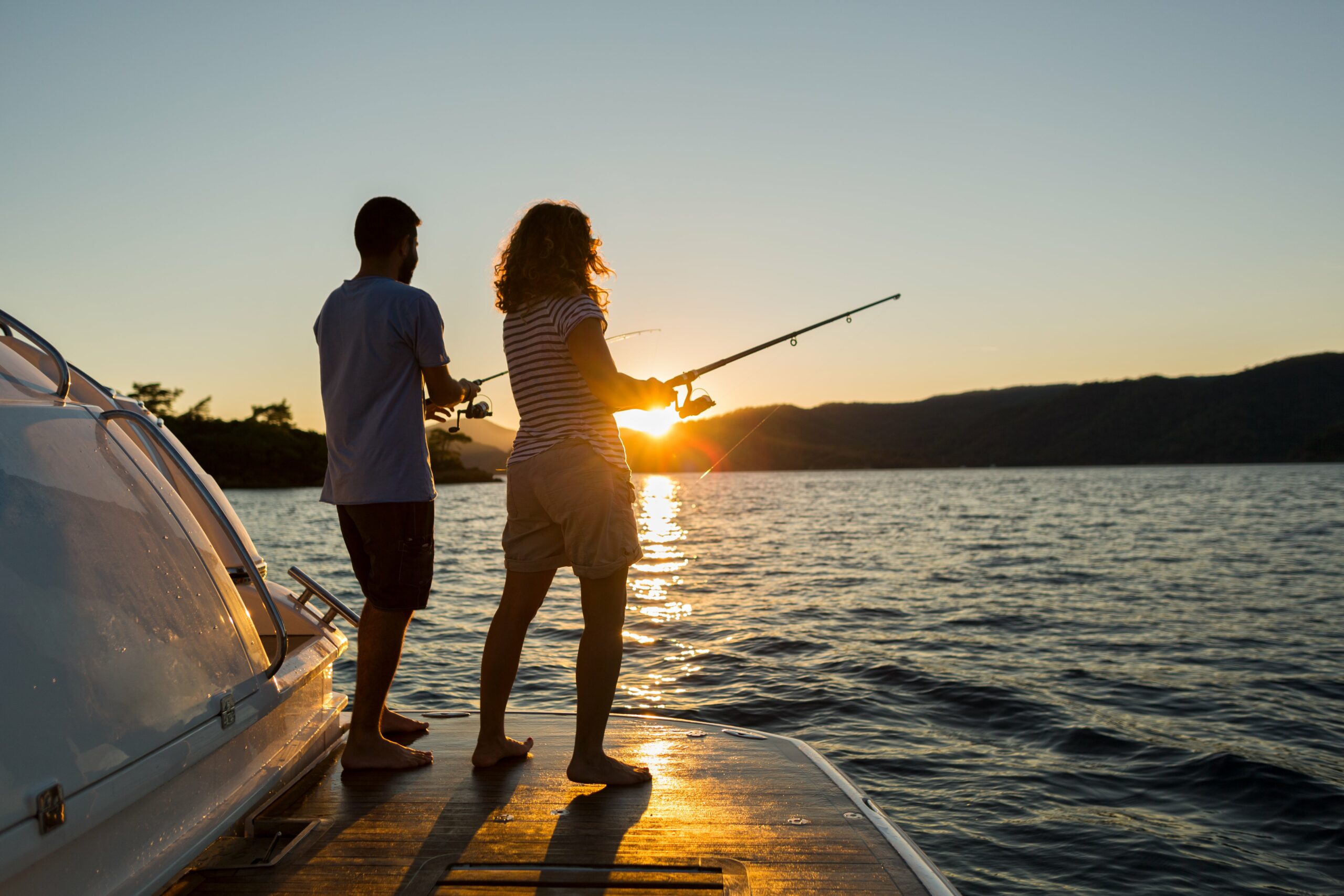 Two people fishing off the edge of a boat during the sunset